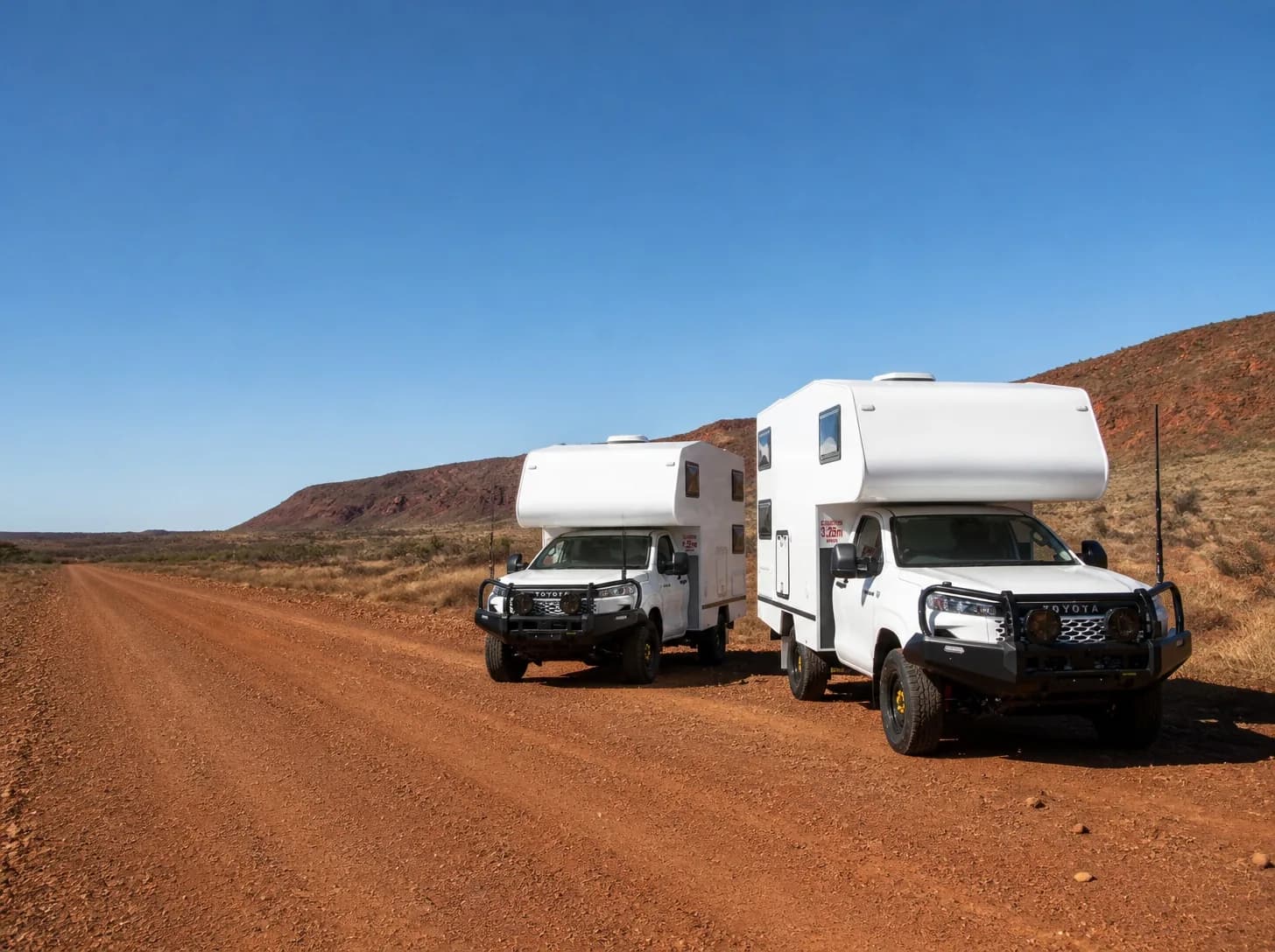 Two matching white living bodies on single-cab utilities, parked on a red dirt road